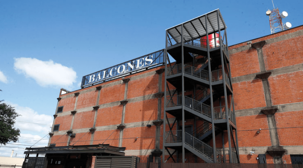 Image of Balcones Distillery. It is a brick building with a metal fire escape. The name appears on the roof in large white lettering. 