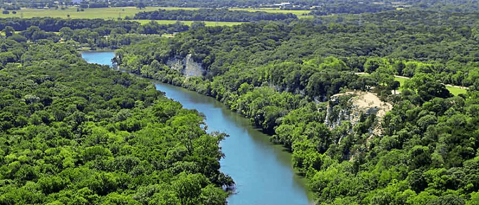 Aerial image of the Brazos River going through Cameron Park. The banks of the river are thick with green tree canopies and two rock faces. 