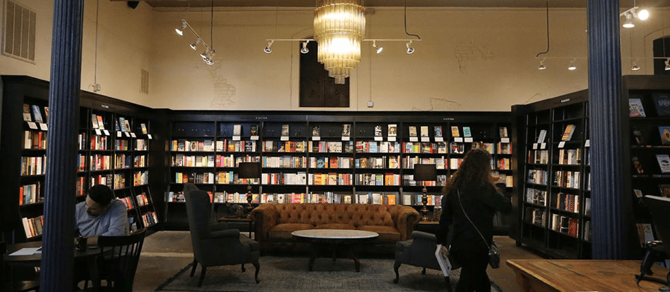 Image of filled bookshelves at Fabled Bookshop in Waco, TX. There are also chairs, tables, and couches, and a chandelier-style light fixture hanging from the ceiling.
